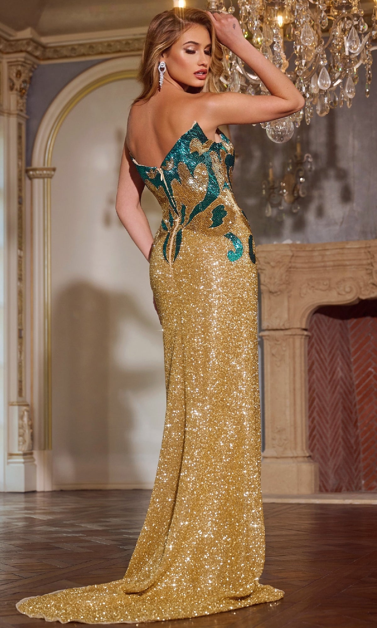 Woman wearing the Jovani D5723 long prom dress, featuring gold sequins, teal floral accents, and a strapless v-neckline, posing elegantly in a room with a chandelier and ornate fireplace.